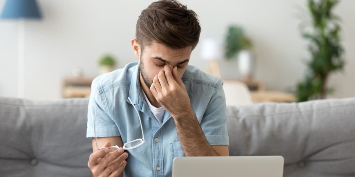 Young man sitting at computer with eyes closed, rubbing eyes due to eye strain.