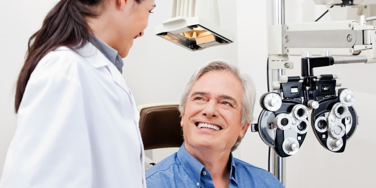 Senior age man sitting in exam chair with phoreopter in the background, talking with eye doctor before having a comprehensive eye exam.