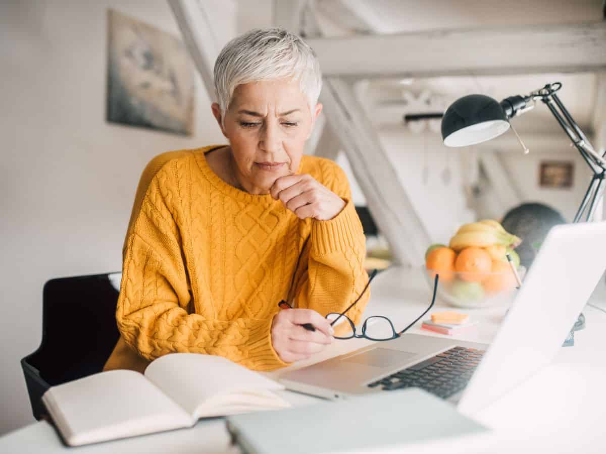 Attractive senior age woman researching information about cataract surgery, reading an open book with an open computer laptop nearby.