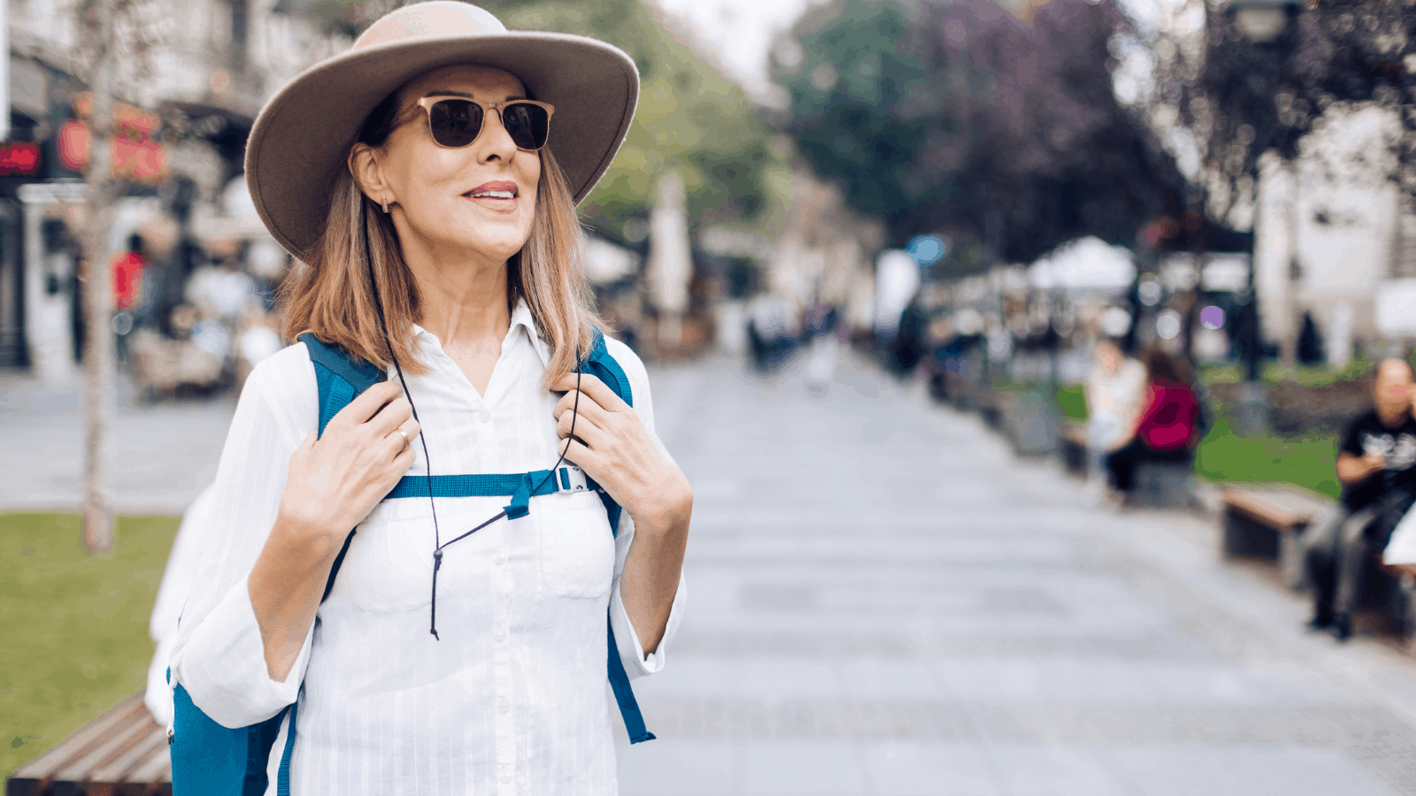 Woman traveling on a city street wearing a backpack, large rim hat, and sunglasses.