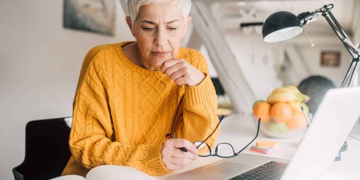 Attractive senior age woman researching information about cataract surgery, reading an open book with an open computer laptop nearby.